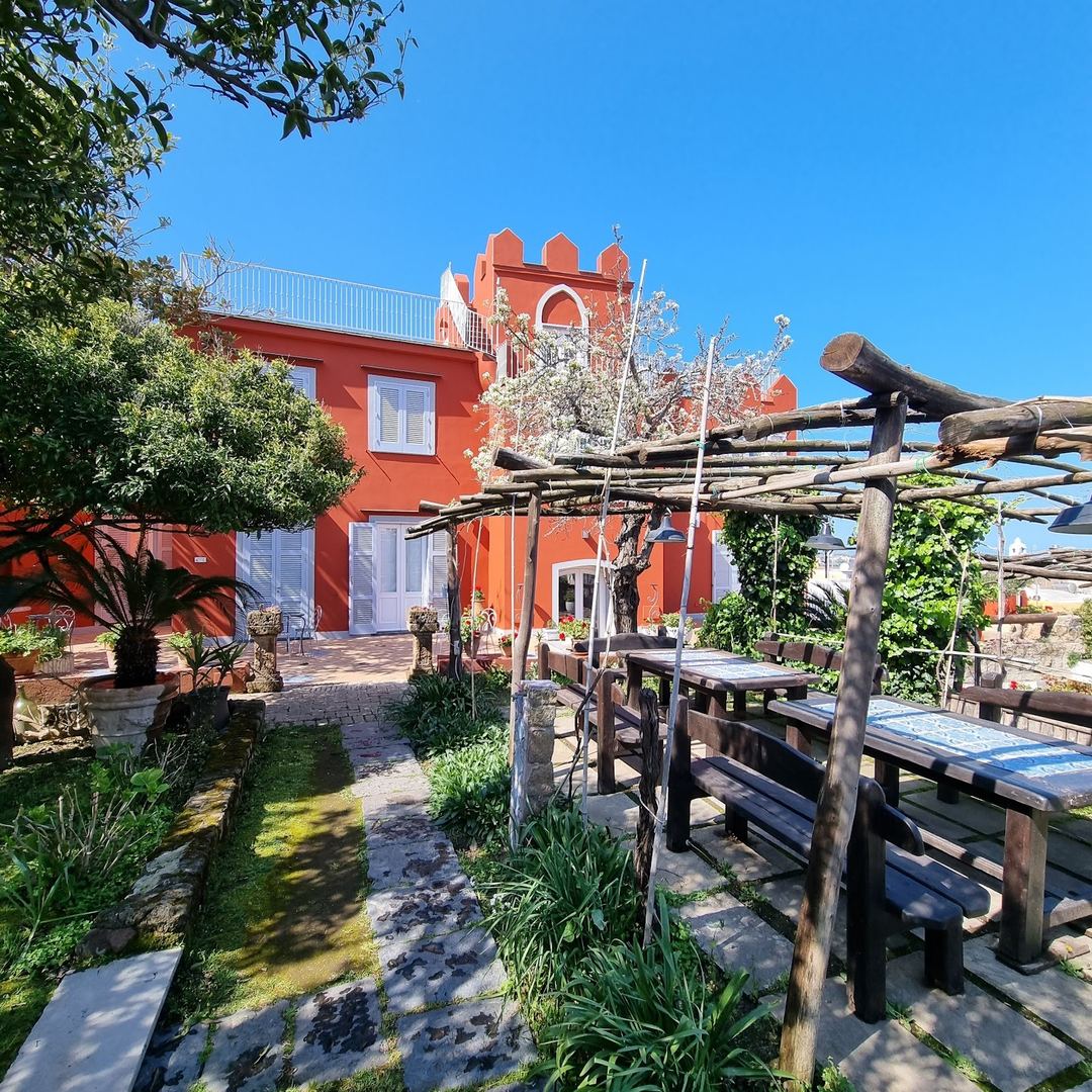 The vibrant red-orange exterior of La Vigna hotel features a rustic wooden pergola with climbing vines and rustic dining tables in the foreground courtyard.