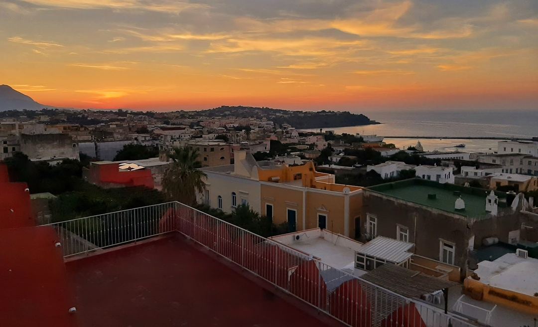 Sunset view from a rooftop terrace overlooking a coastal Mediterranean town with colorful buildings and the sea in the distance.