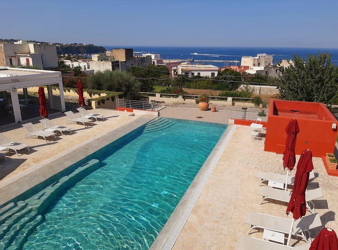 The hotel's rectangular swimming pool with turquoise water, surrounded by loungers with red umbrellas, overlooking the Mediterranean coastline.