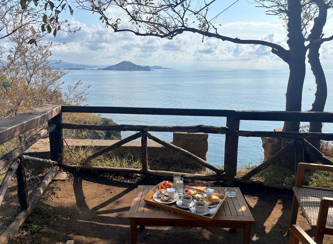 A seaside breakfast setup on a wooden terrace overlooking a calm blue sea with distant islands and mountains under partly cloudy skies.
